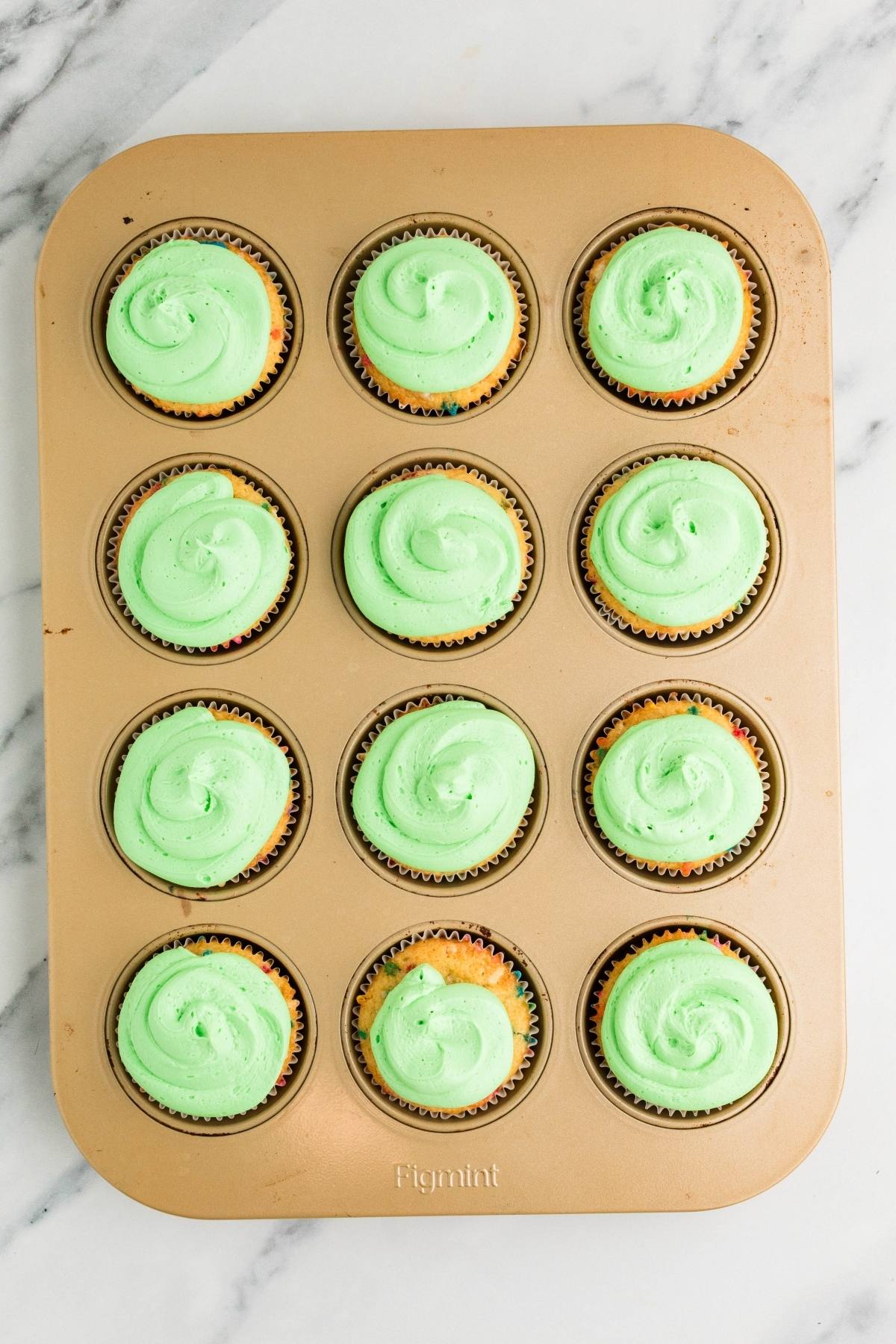 A muffin tin with twelve cupcakes topped with light green swirled frosting on a marble surface.