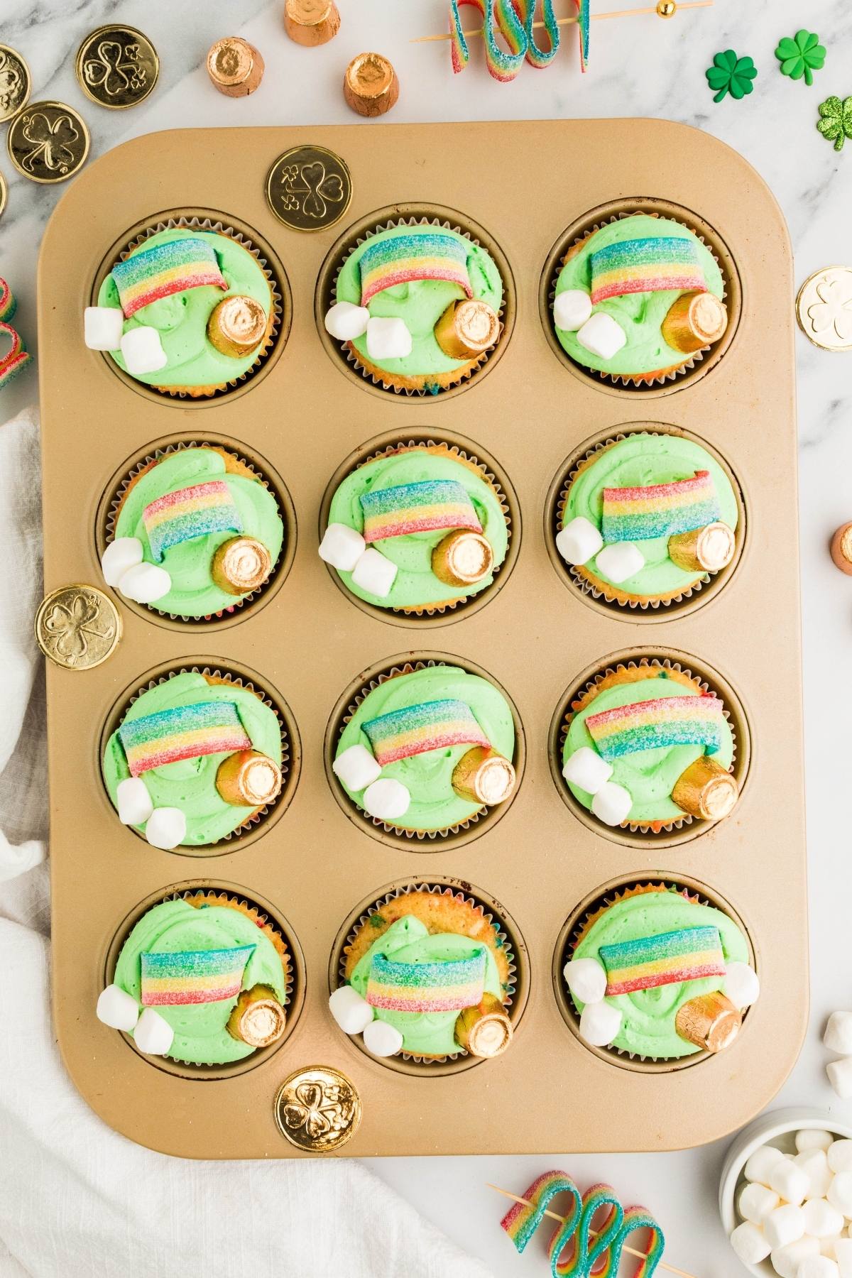 A muffin tin with 12 cupcakes topped with green frosting, gold coins, rainbows, and mini marshmallows.