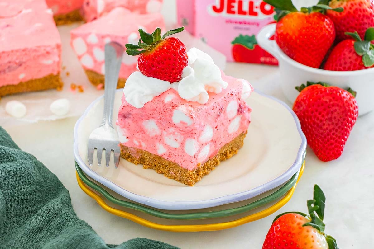 A slice of pink strawberry chiffon squares dessert with marshmallows, whipped cream, and a strawberry on top, served on a stacked plate.