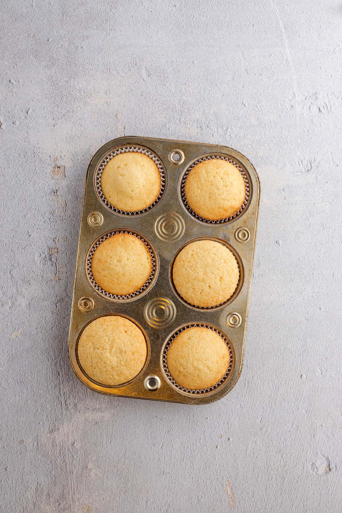 Six plain Strawberry Shortcake Cupcakes in a metal muffin tin, viewed from above on a light gray surface.