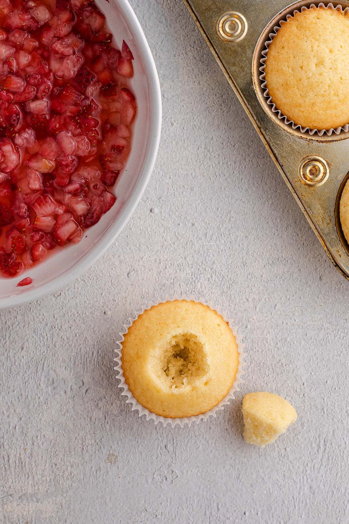 A plain Strawberry Shortcake Cupcake with its center removed sits beside the extracted piece; strawberry filling and more cupcakes are nearby.
