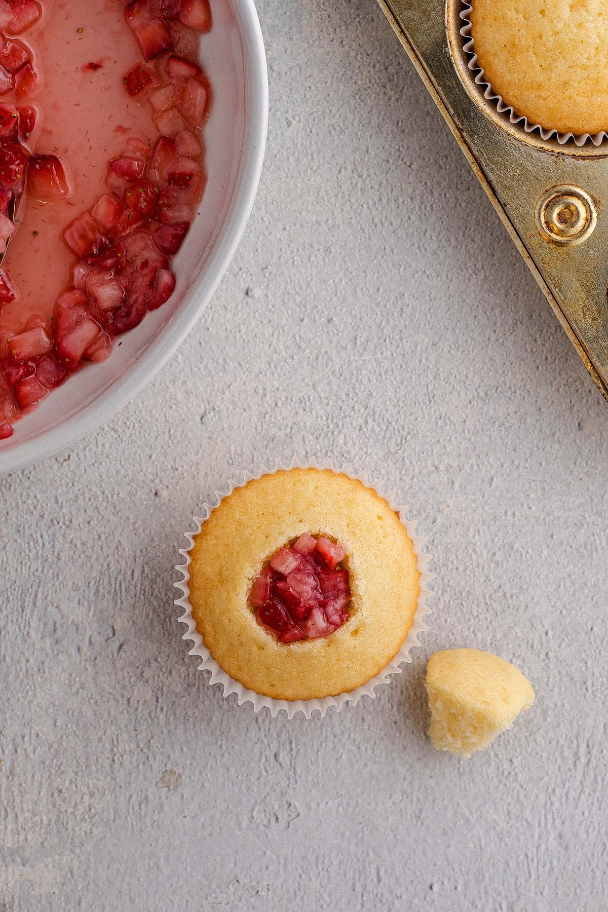 Strawberry Shortcake Cupcakes with a piece removed, filled with strawberry compote, sit beside a muffin tin and a bowl of compote.