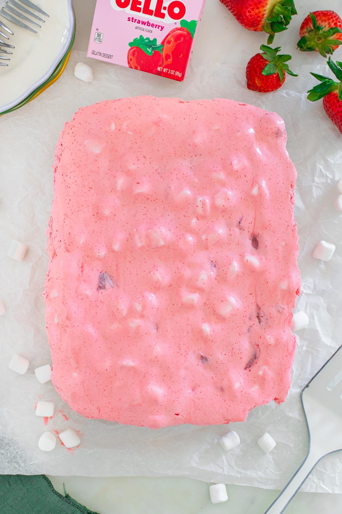 A pink strawberry Jello dessert with marshmallows on parchment paper, with fresh strawberries nearby.