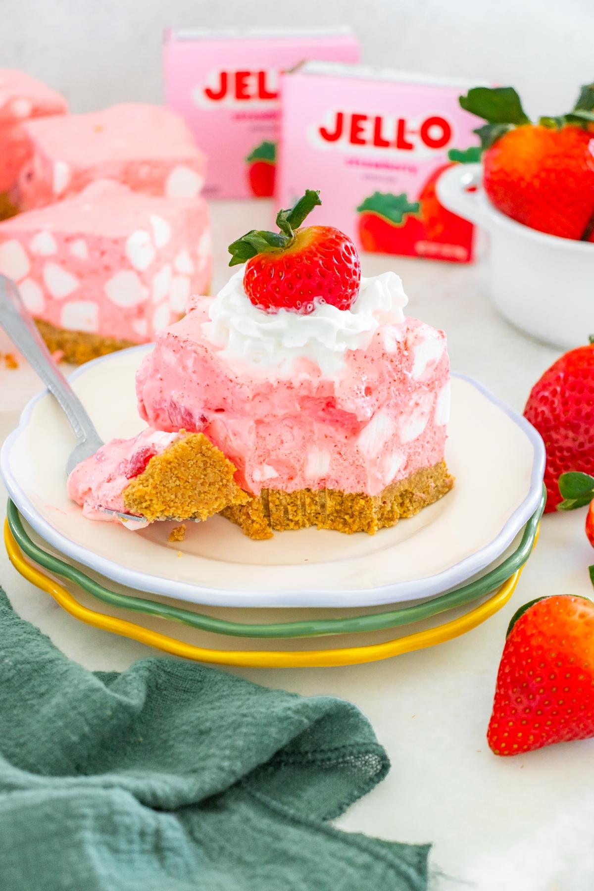 A slice of strawberry chiffon squares dessert with whipped cream and a strawberry on top, sitting on a plate.