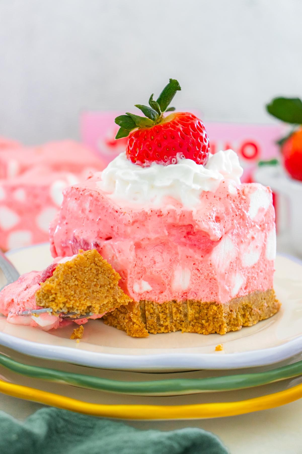 A slice of strawberry chiffon squares dessert with a graham cracker crust, whipped cream, and a whole strawberry on top.