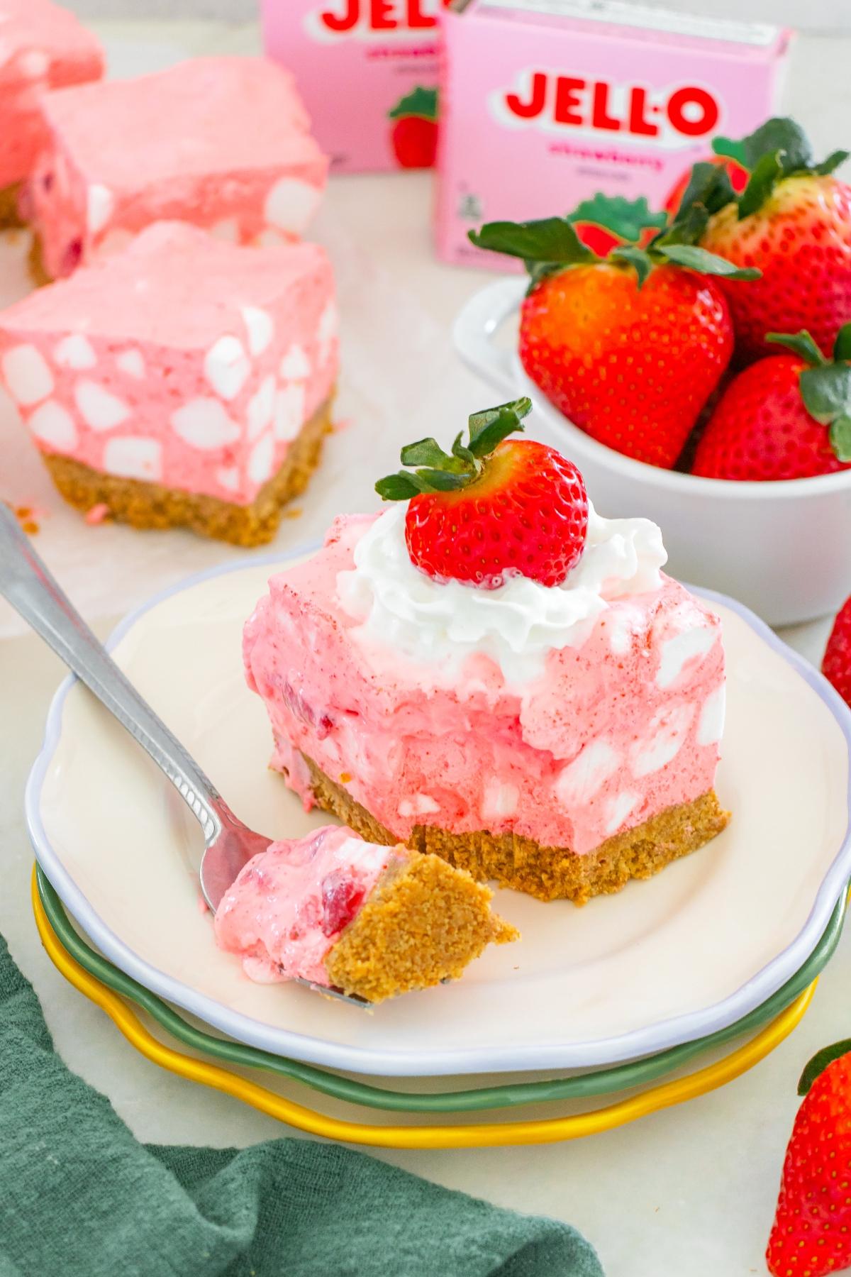 A slice of strawberry chiffon squares dessert with whipped cream and a strawberry on top, next to fresh strawberries.