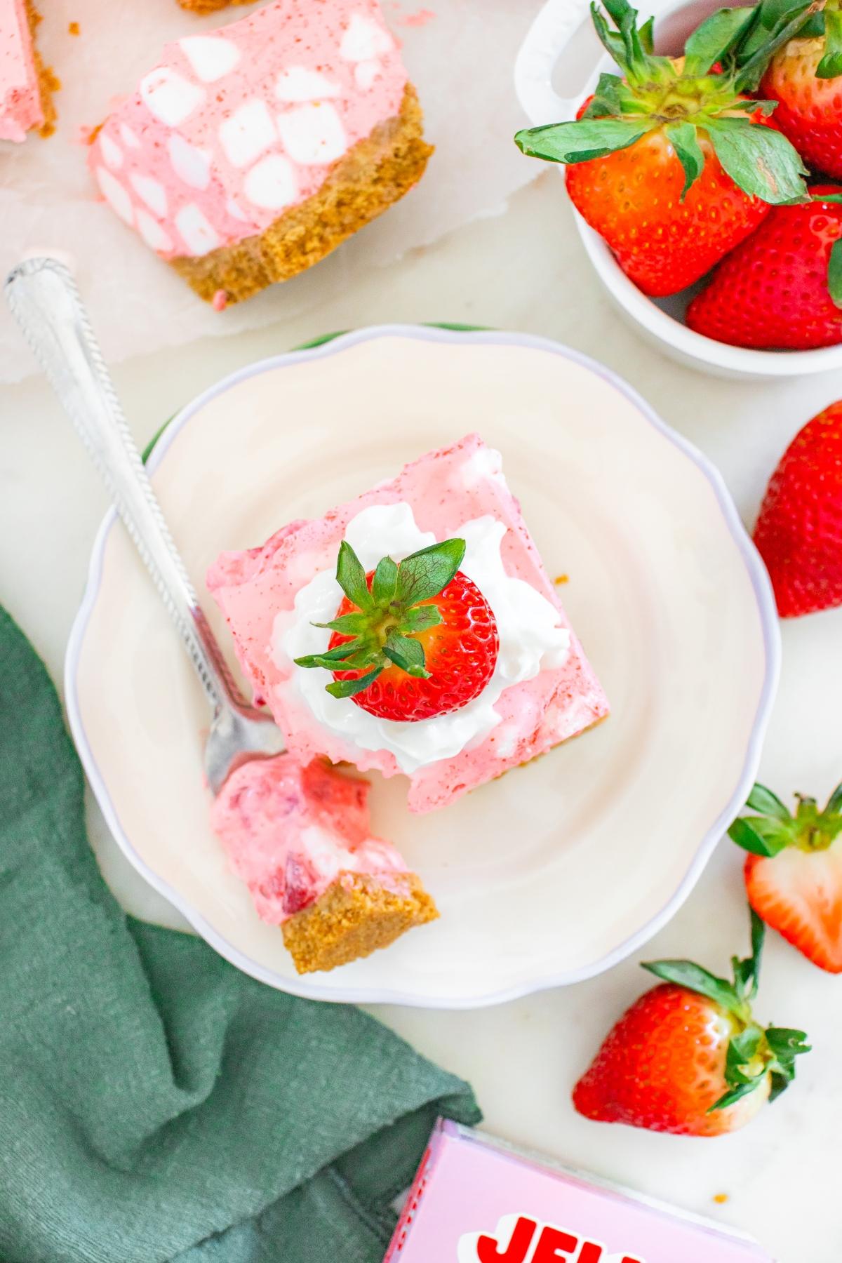 A slice of strawberry chiffon squares dessert with whipped cream and a strawberry on top, on a plate with a fork.