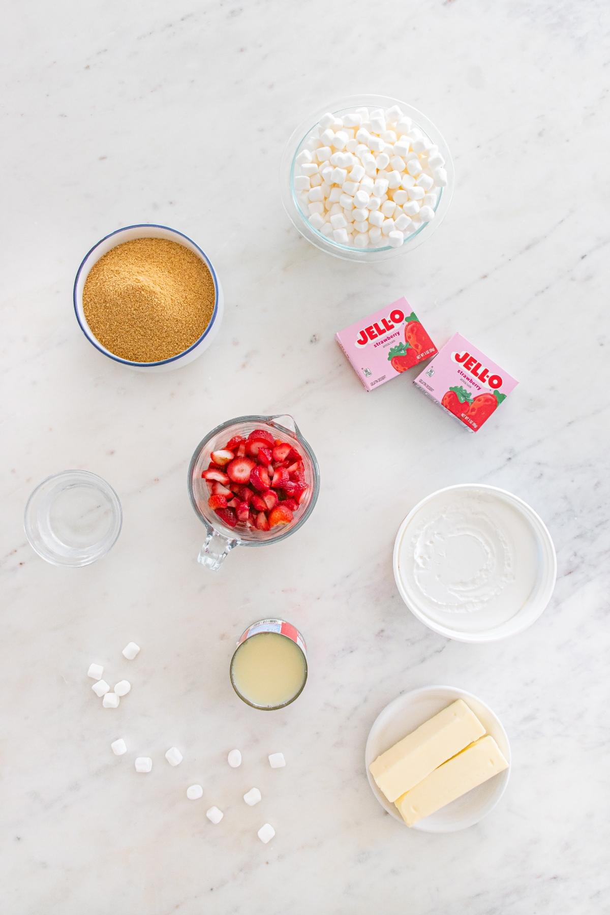 Ingredients for a dessert: marshmallows, graham crumbs, strawberries, Jell-O, butter, cream, and condensed milk.
