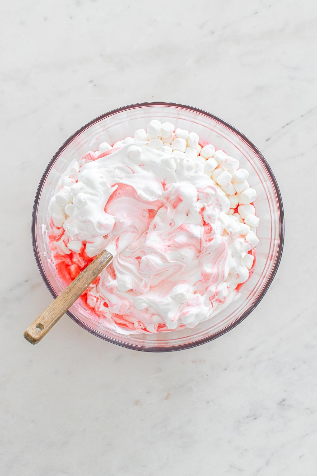 A glass bowl with white and pink swirled whipped cream, mini marshmallows, and a wooden spatula.