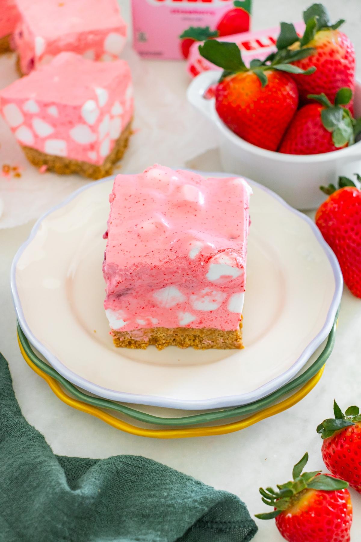 A pink strawberry chiffon squares dessert bar with marshmallows on a plate, next to fresh strawberries.