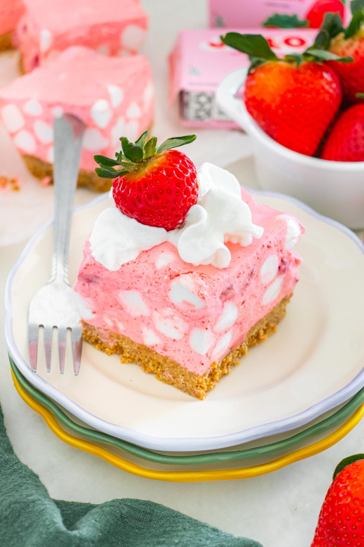 A slice of pink strawberry chiffon squares dessert with marshmallows, whipped cream, and a fresh strawberry on top.