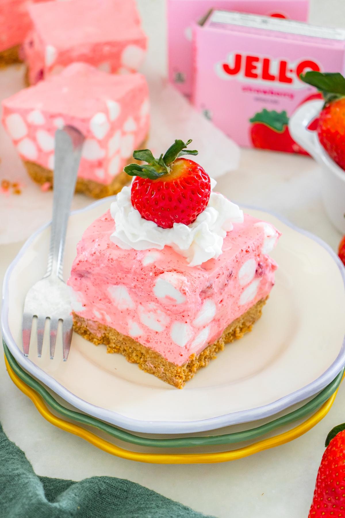 A pink strawberry chiffon squares dessert with marshmallows, whipped cream, and a strawberry on top, served on a plate.
