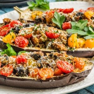 Three baked stuffed eggplant halves filled with tomatoes, olives, herbs, and cheese are served on a white plate.