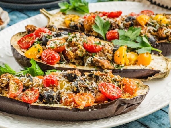 Three baked stuffed eggplant halves filled with tomatoes, olives, herbs, and cheese are served on a white plate.
