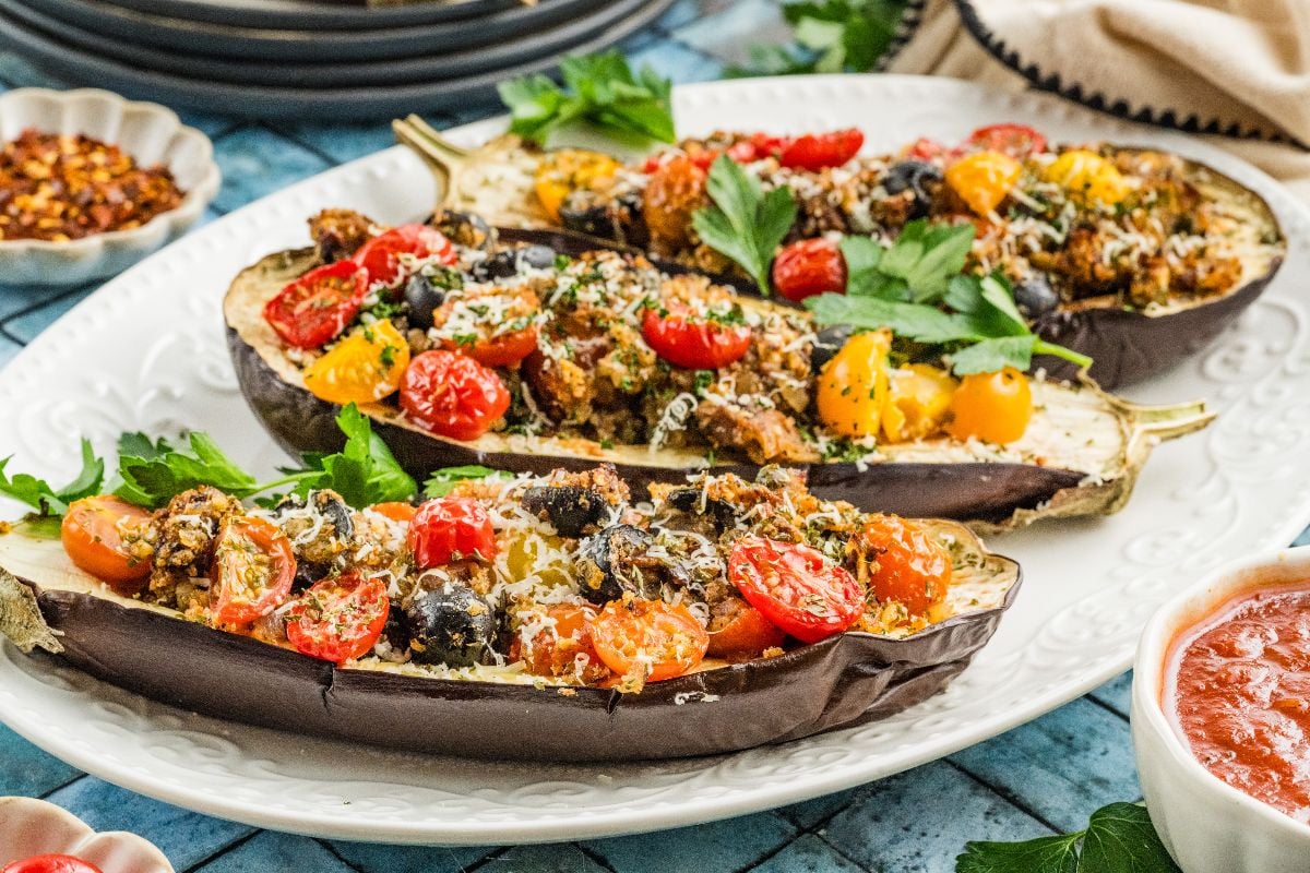 Halved stuffed eggplant filled with tomatoes, olives, herbs, and cheese, served on a white plate with a parsley garnish.