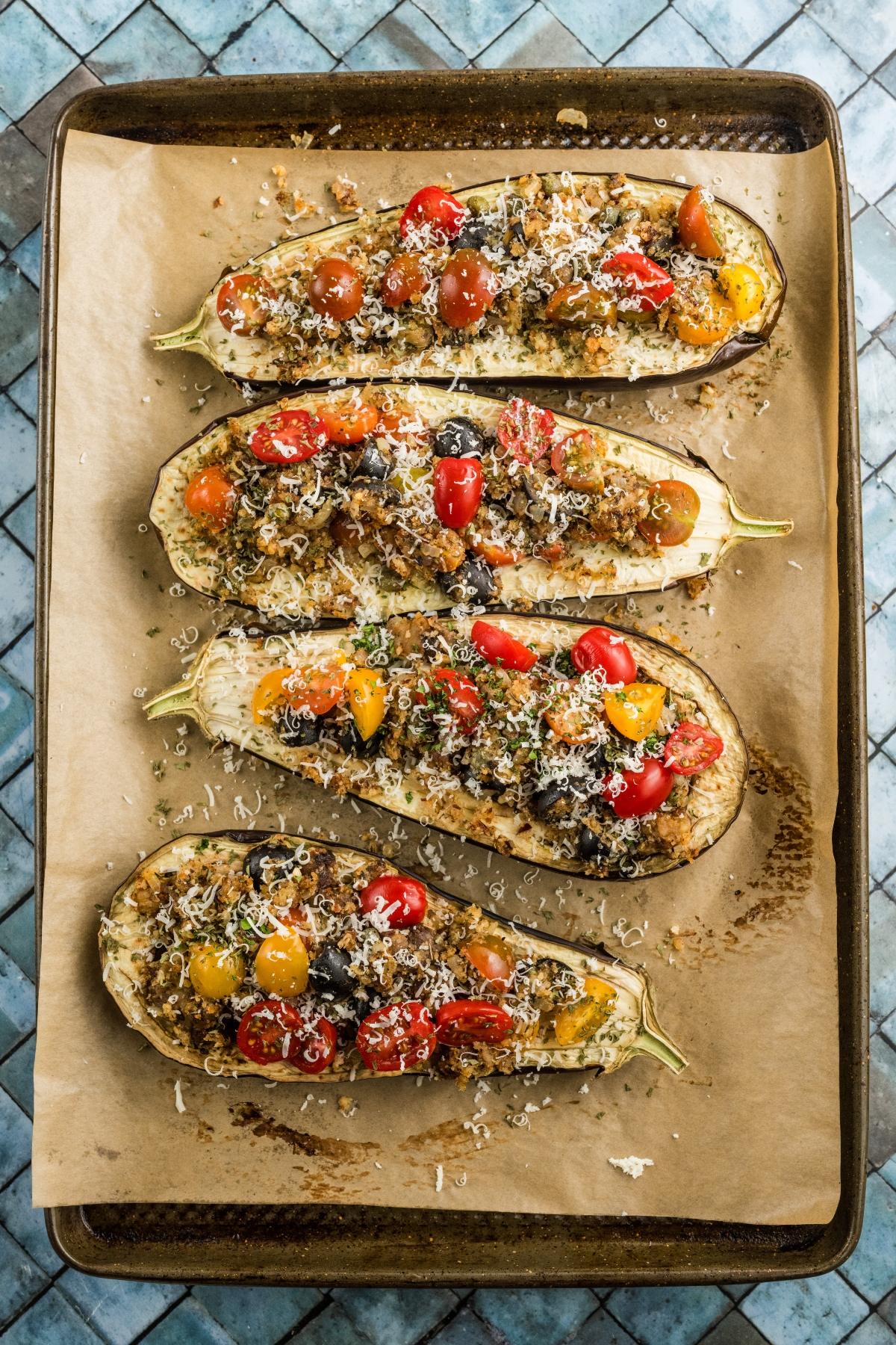 Four stuffed eggplant halves topped with cherry tomatoes and grated cheese on a parchment-lined baking sheet.