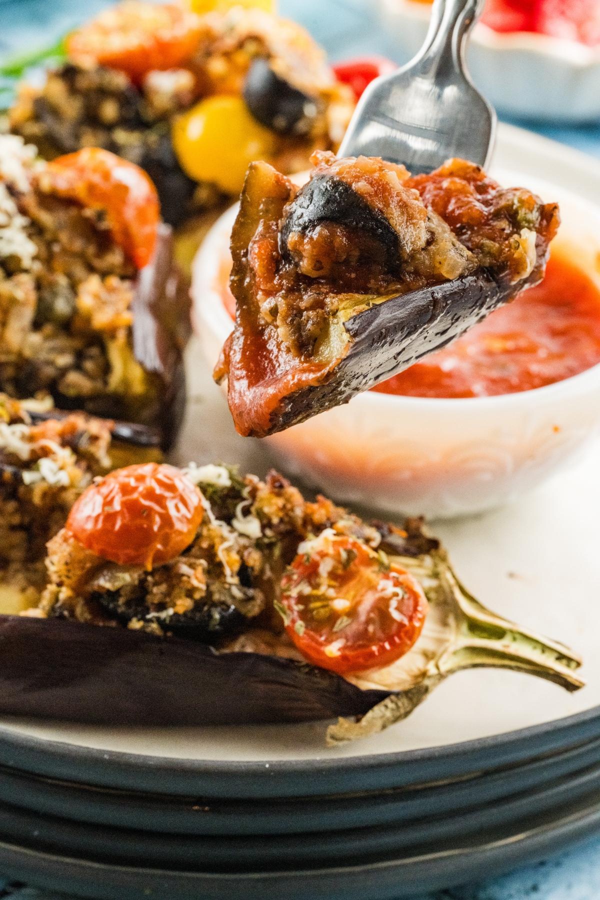 A fork holds a bite of stuffed eggplant with tomatoes and sauce, with more stuffed eggplant on a plate.
