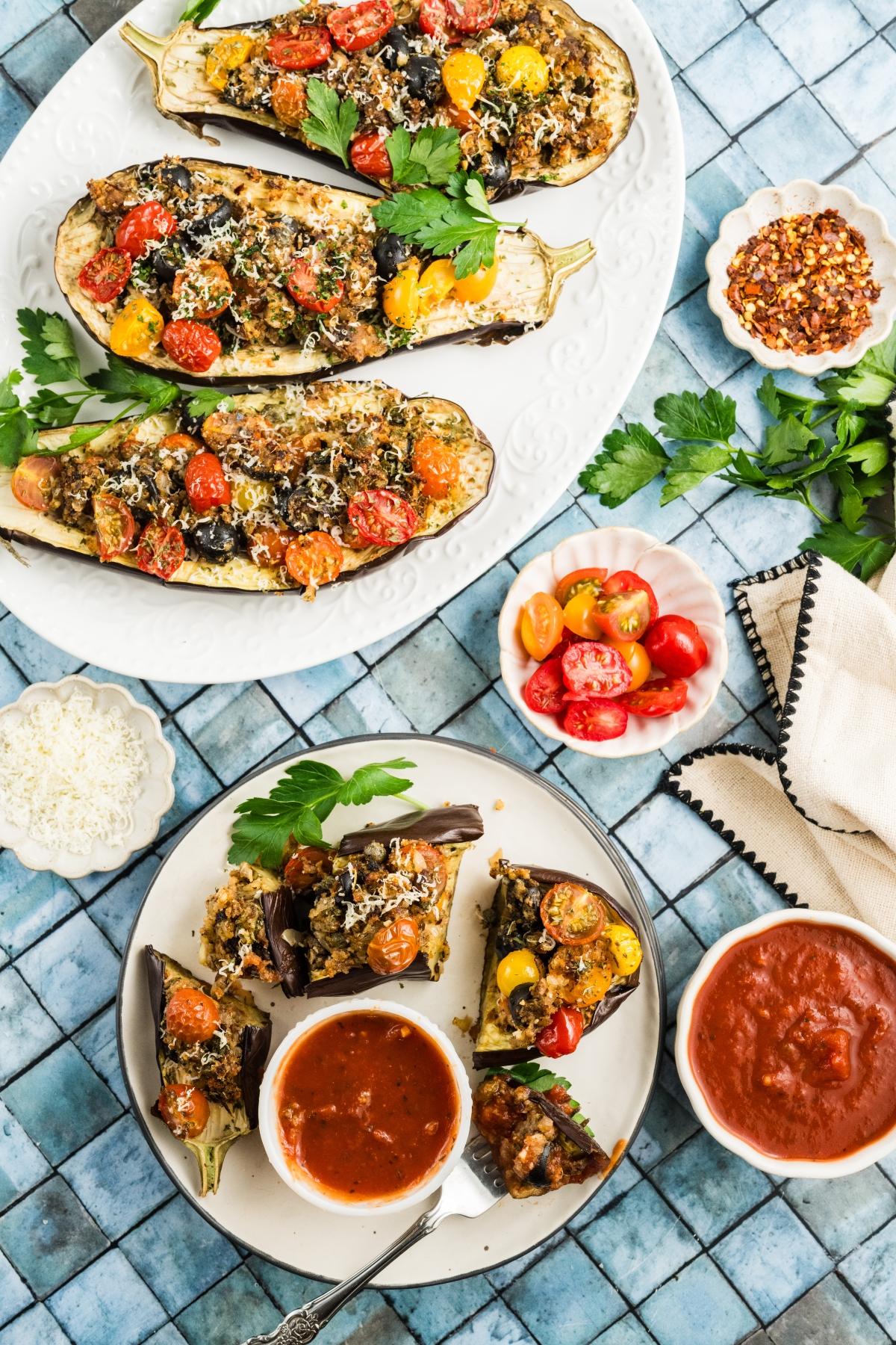Stuffed eggplant halves with tomatoes and herbs on plates, served with tomato sauce and garnishes.