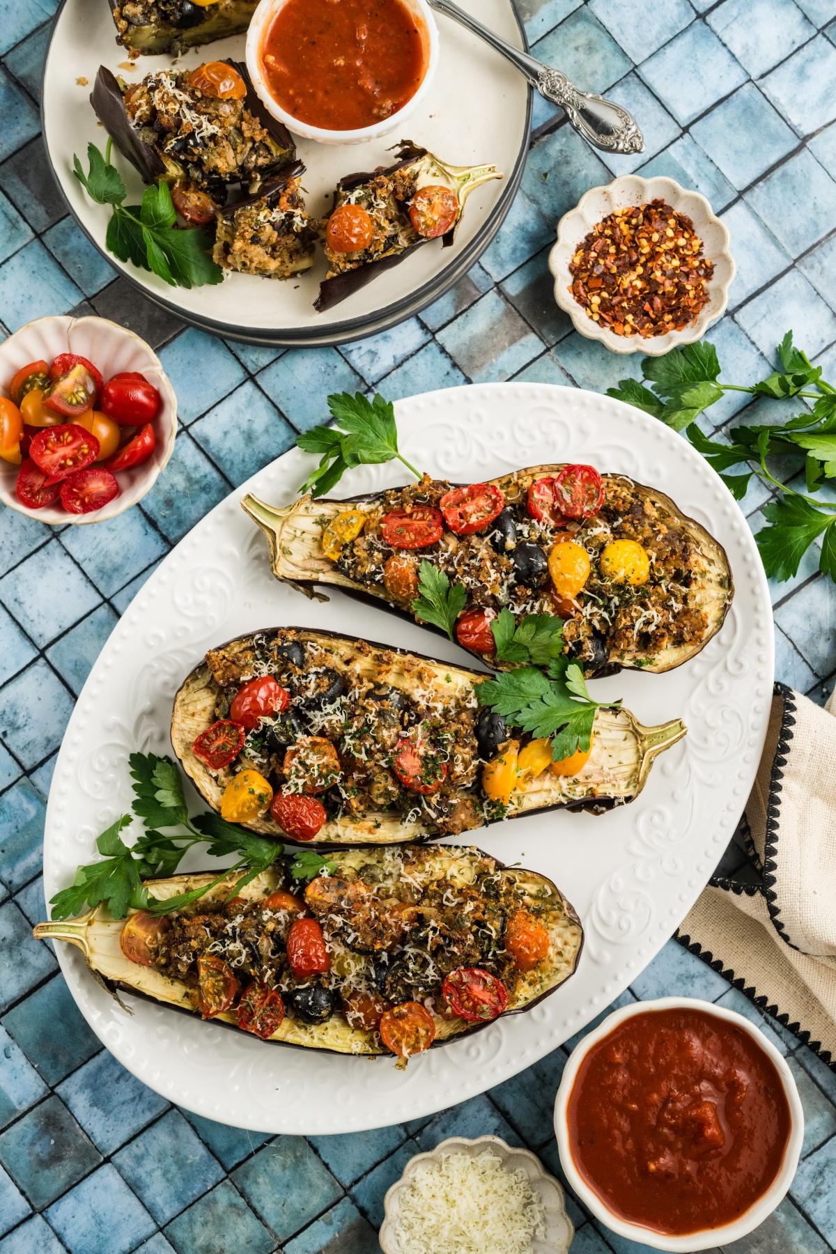 Stuffed eggplant halves topped with tomatoes and herbs on a white platter, with sauces and parsley on the side.