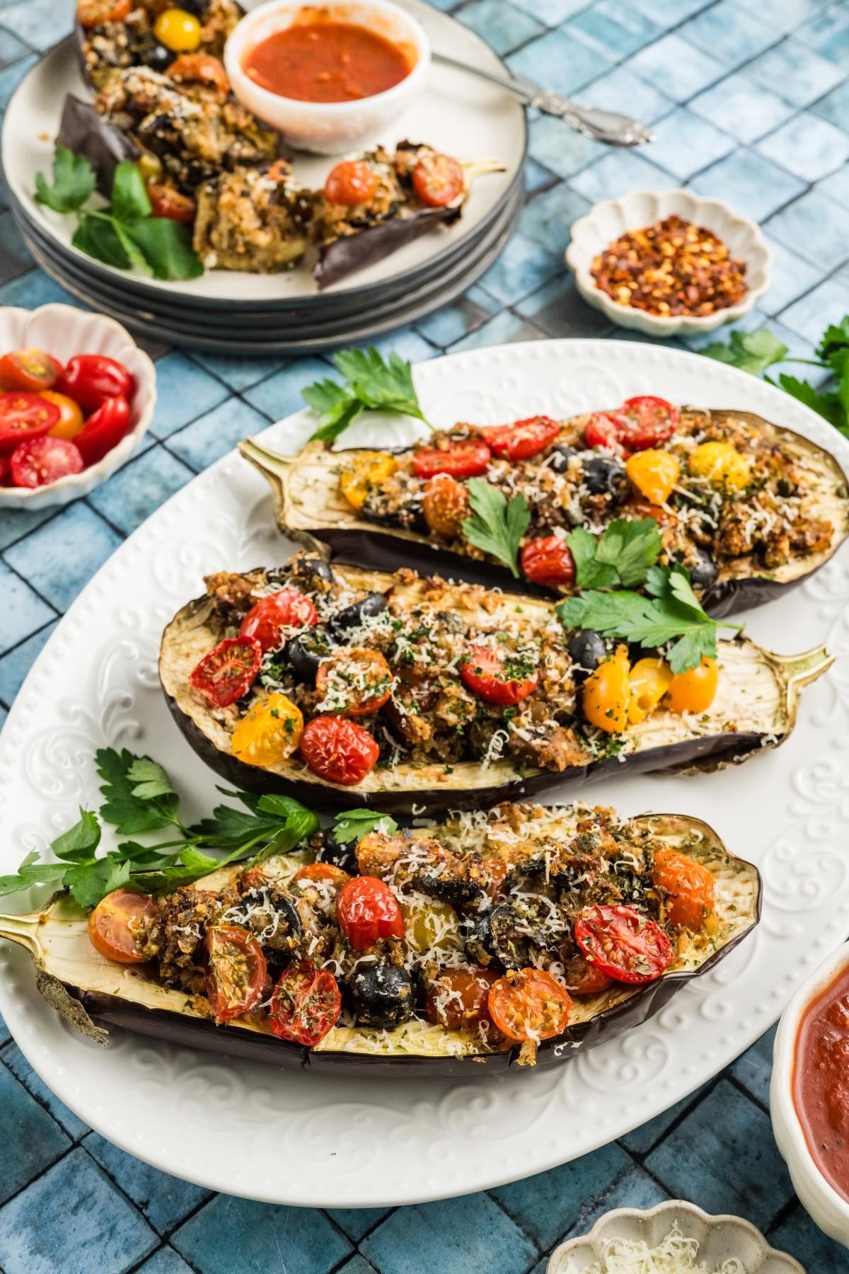 Three stuffed eggplant halves topped with tomatoes and herbs on a white platter, with sauce and garnishes nearby.