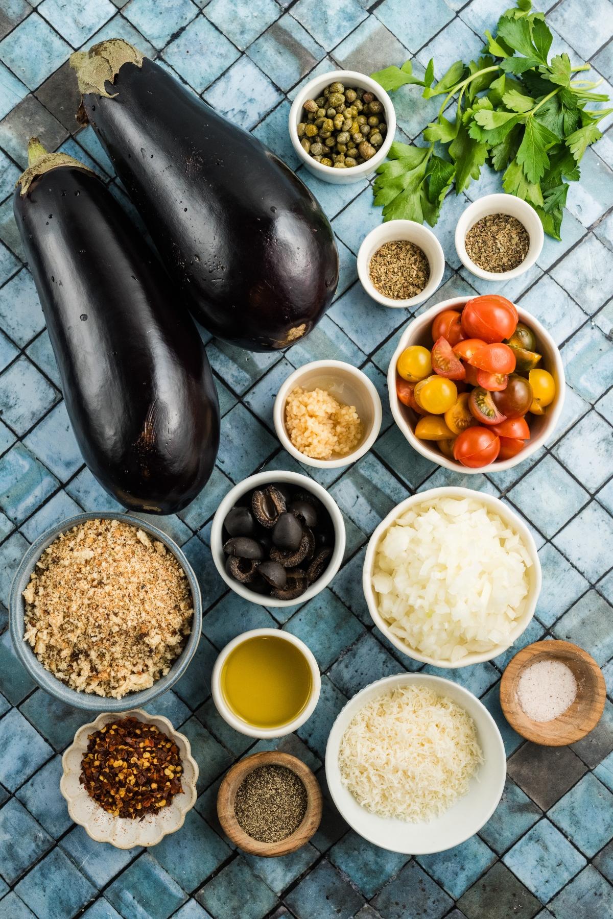 Ingredients for a dish, including eggplants, tomatoes, onions, olives, oil, spices, and herbs on a blue tile surface.