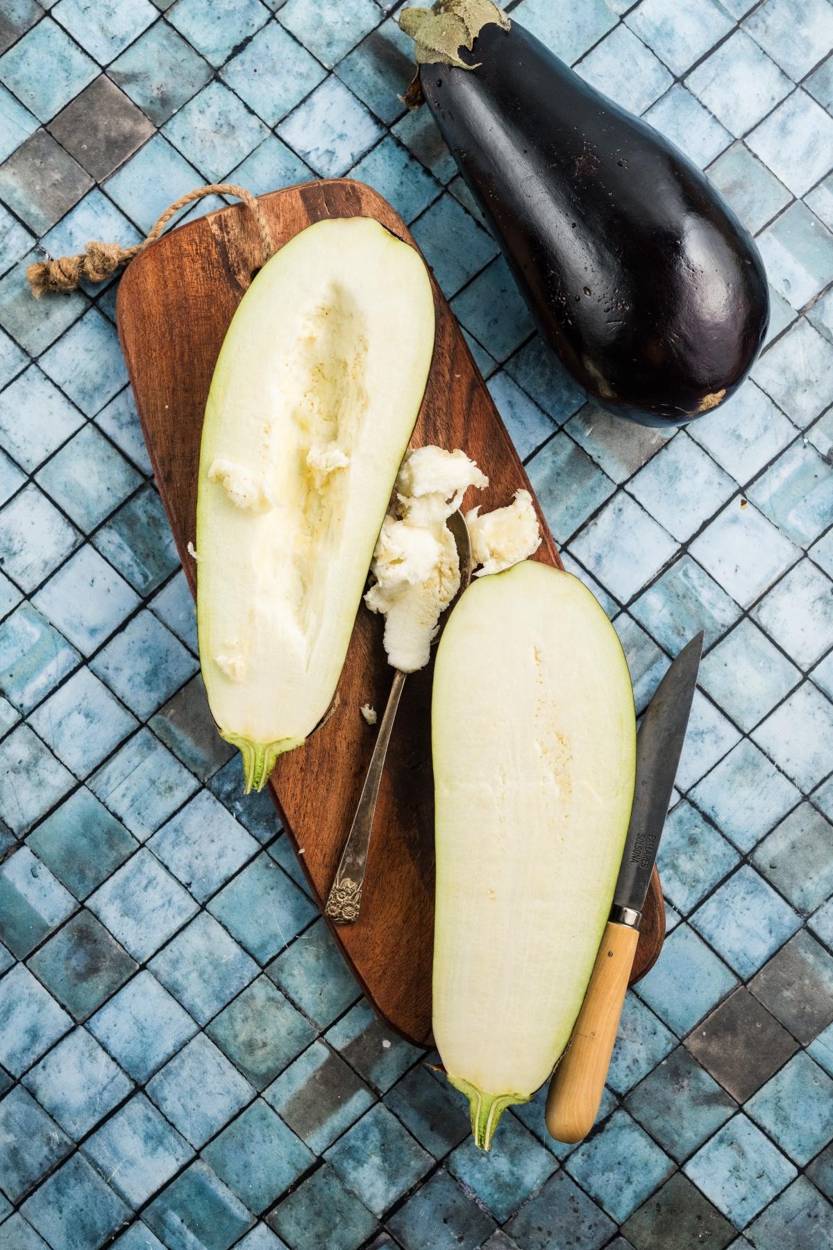 A whole eggplant and a halved eggplant on a wooden board with a knife on blue tile.