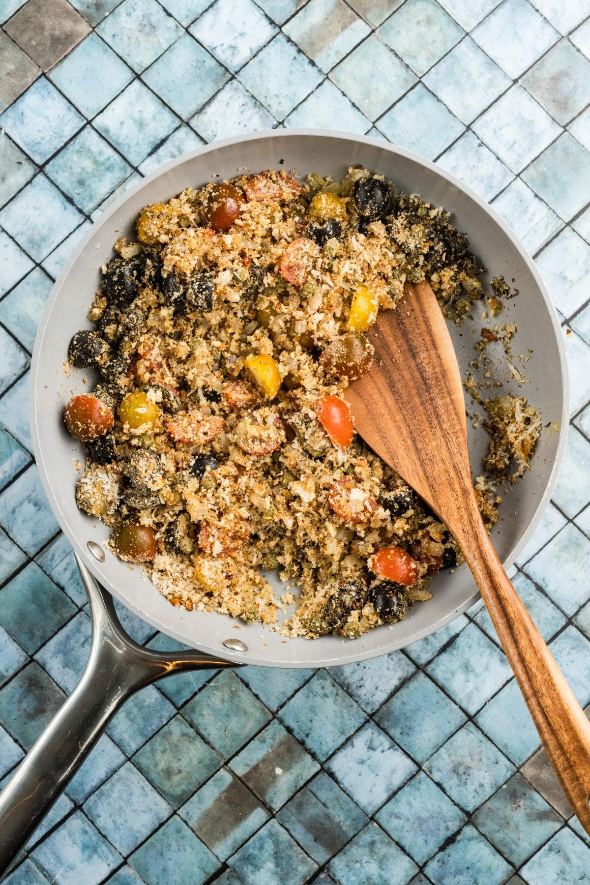 A skillet with breadcrumbs, cherry tomatoes, and black olives, stirred with a wooden spatula on a tiled surface.