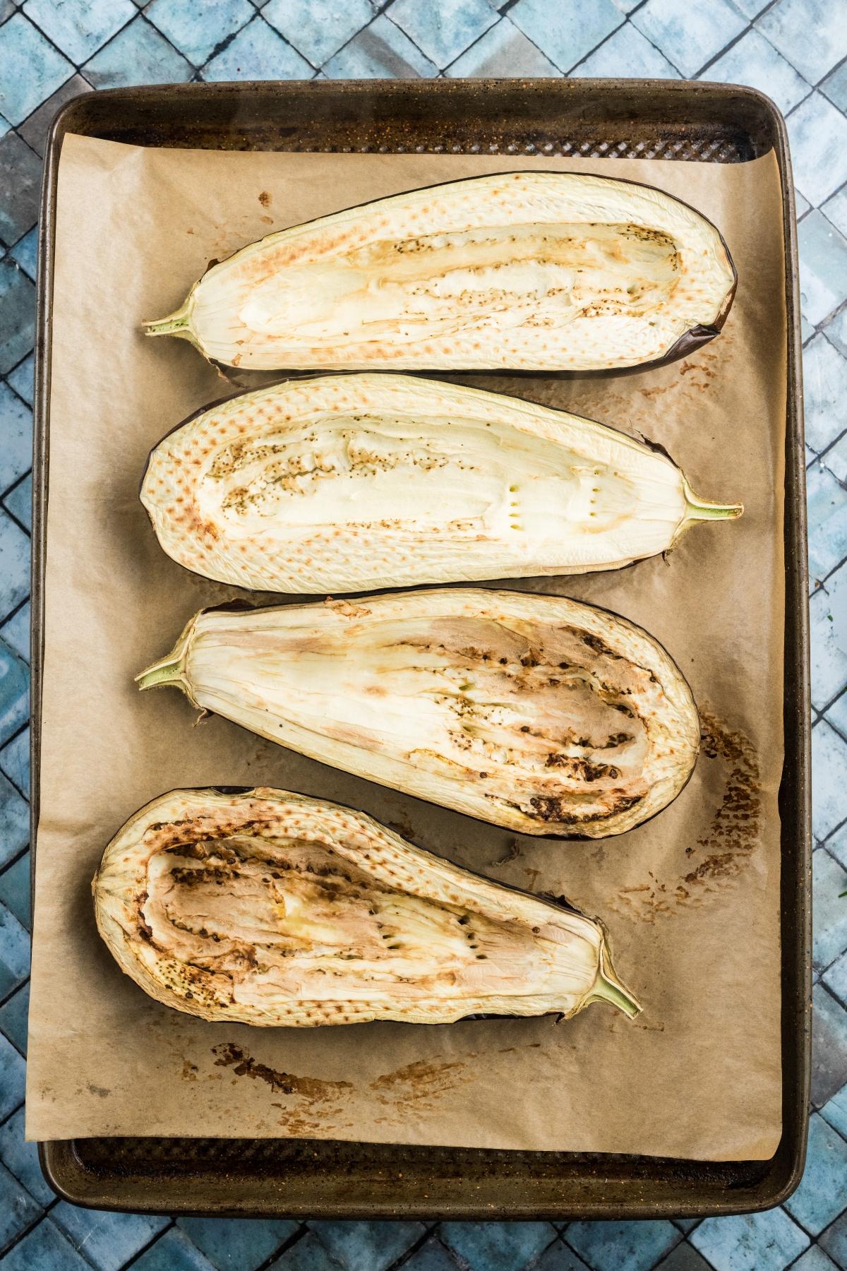 Four halved eggplants, scored and roasted, on parchment-lined baking sheet atop a blue tiled surface.