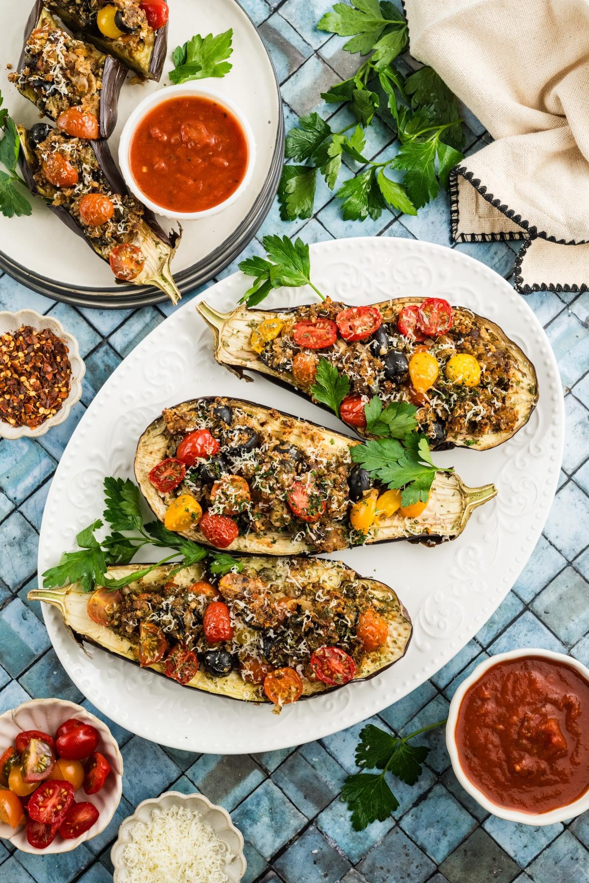 Stuffed eggplants with tomatoes, herbs, and cheese on a platter, with bowls of sauce and seasonings nearby.
