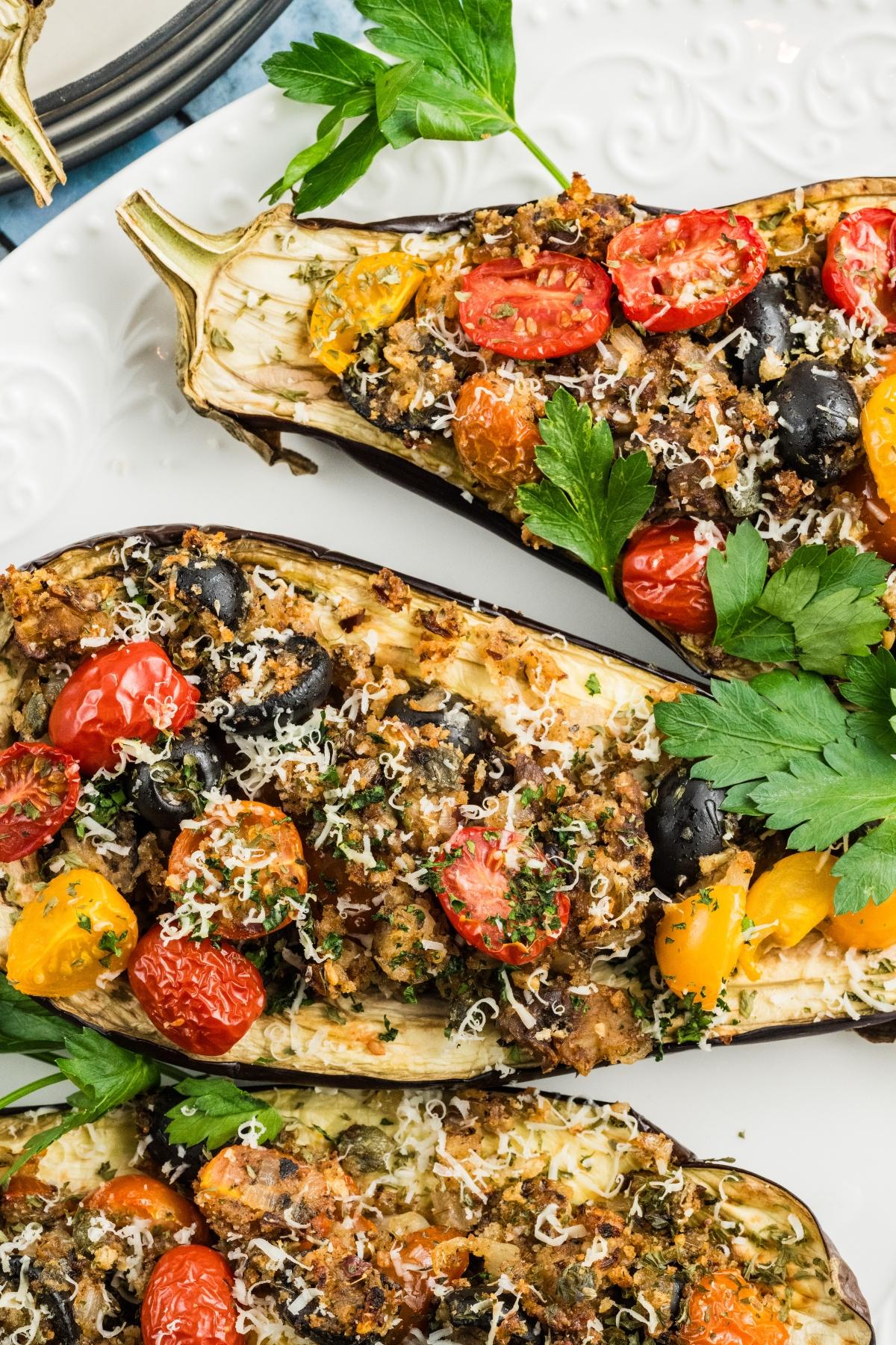 Halved eggplants stuffed with tomatoes, olives, herbs, and cheese, garnished with fresh parsley on a white plate.