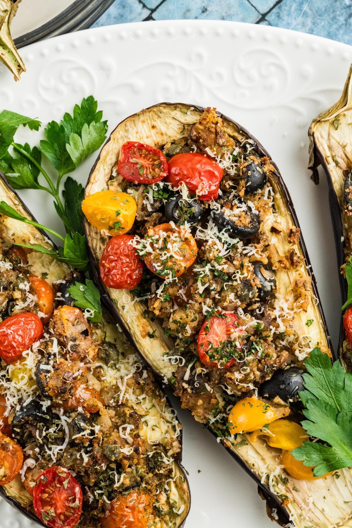 Roasted stuffed eggplant with cherry tomatoes, black olives, herbs, and grated cheese on a white plate.