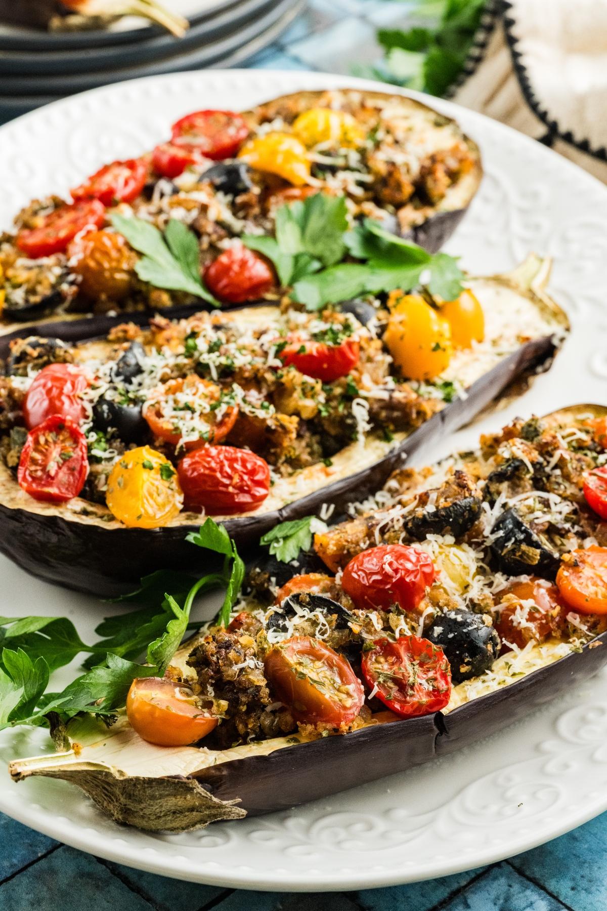 Stuffed eggplant halves with tomatoes, olives, herbs, and cheese on a white plate, garnished with parsley.