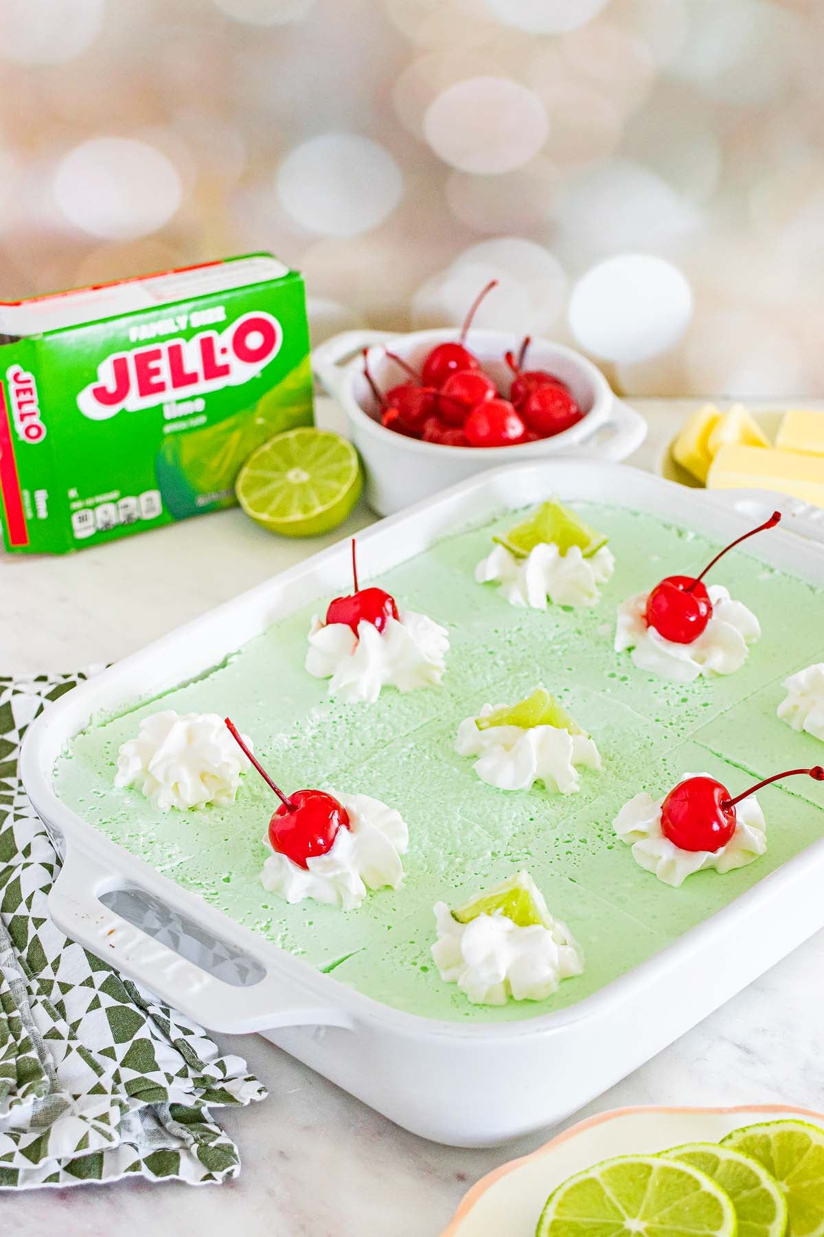 A classic Lime Jello Salad dessert, topped with whipped cream and cherries in a white dish, with fresh limes and a box of Jell-O nearby.