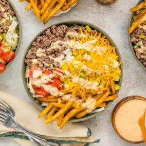 Big Mac Salad bowls with ground beef, fries, cheese, tomatoes, pickles, and special sauce on a table, served with utensils and extra dipping sauce.
