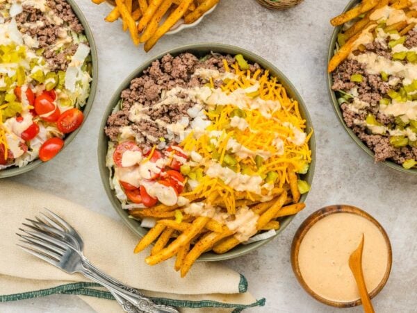 Big Mac Salad bowls with ground beef, fries, cheese, tomatoes, pickles, and special sauce on a table, served with utensils and extra dipping sauce.