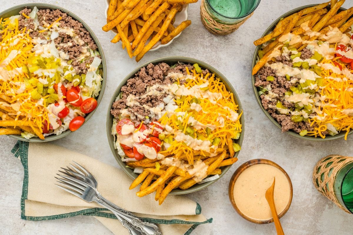 Three bowls of seasoned fries topped with ground beef, cheese, pickles, tomatoes, and creamy sauce.