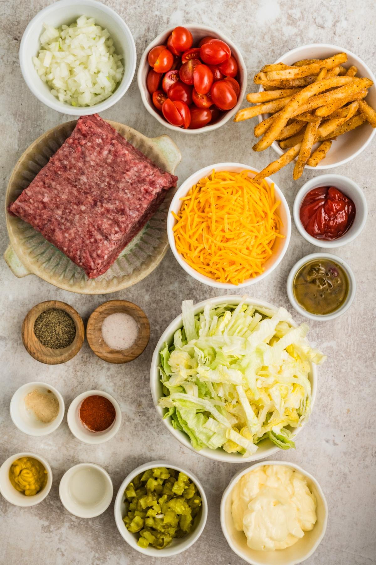 Burger bowl ingredients: ground beef, shredded cheddar, fries, lettuce, tomatoes, sauces, onions, and seasonings.
