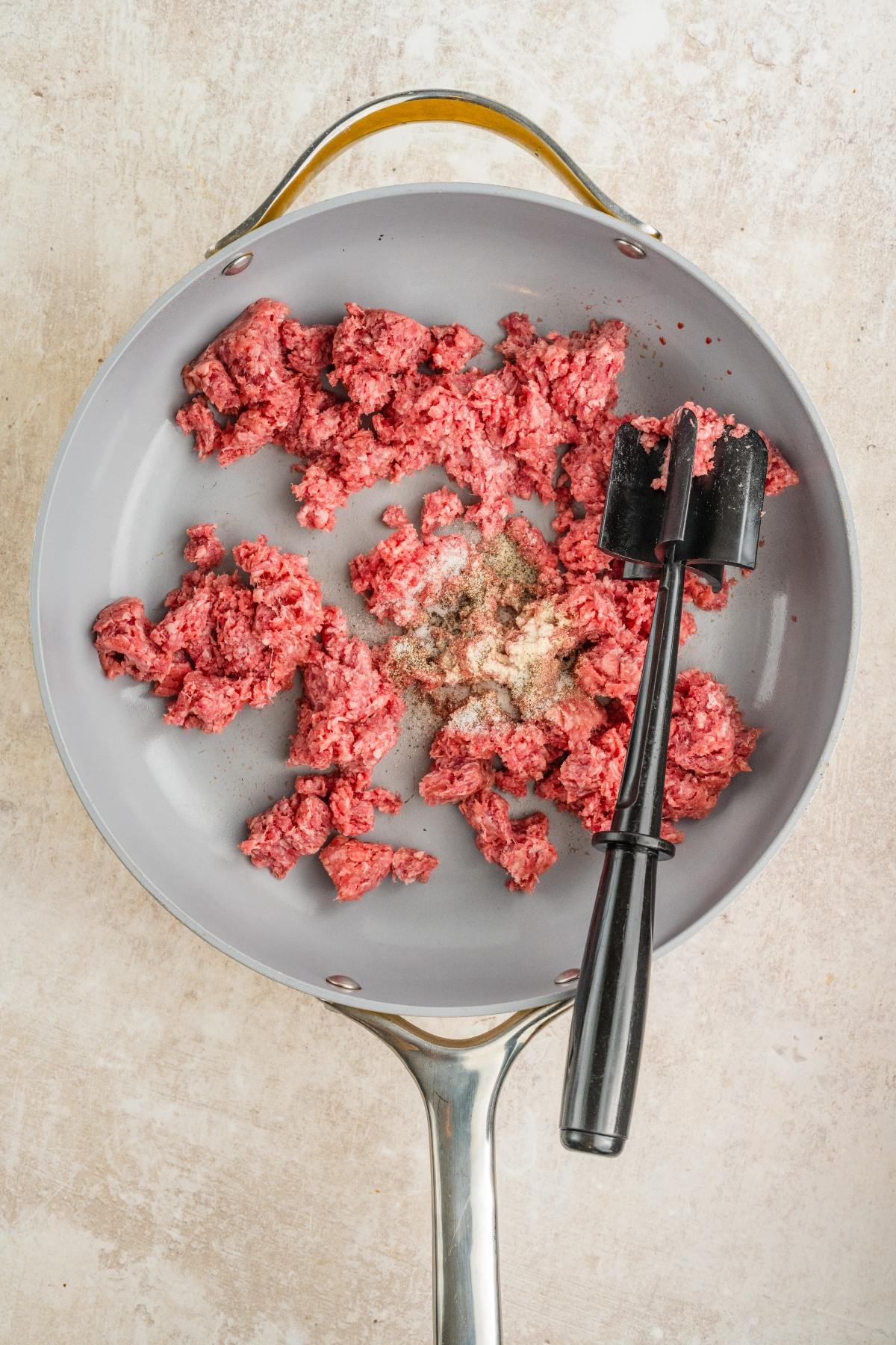 Raw ground beef with seasoning and a black meat chopper in a gray skillet on a light surface.