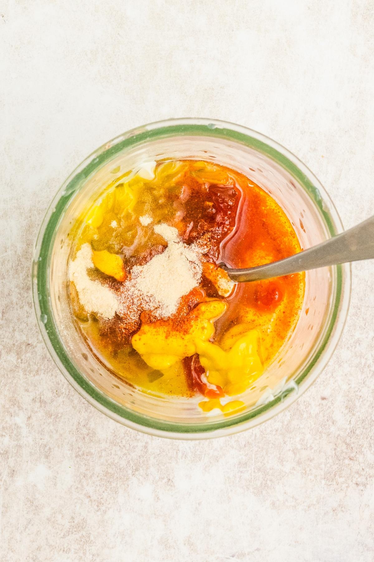 Glass jar with mustard, ketchup, spices, and oil, with a spoon inside, on a light surface.
