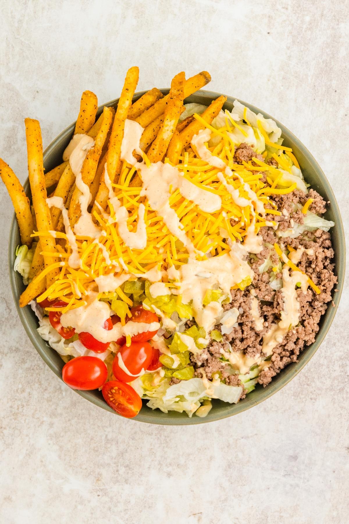 A salad bowl topped with ground beef, shredded cheese, fries, tomatoes, pickles, and creamy dressing.