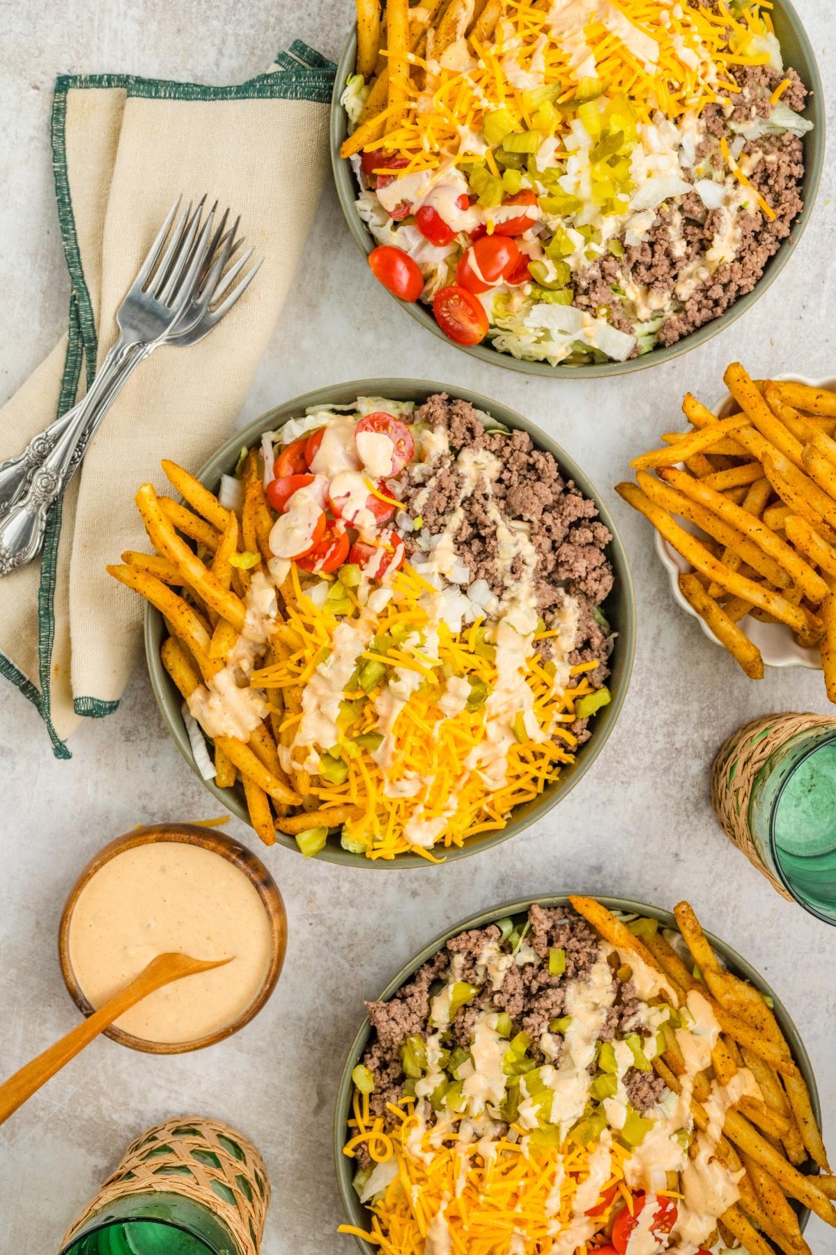 Three bowls of big mac salad with fries, cheese, tomatoes, lettuce, and creamy dressing, with drinks nearby.