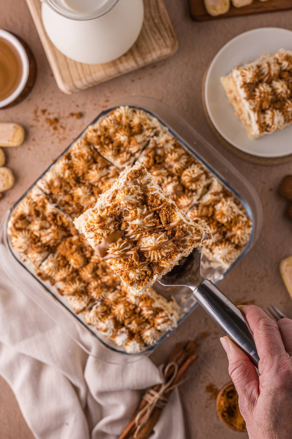 Hand holding a spatula with a square of Biscoff tiramisu over a dish, topped with whipped cream and cocoa powder.
