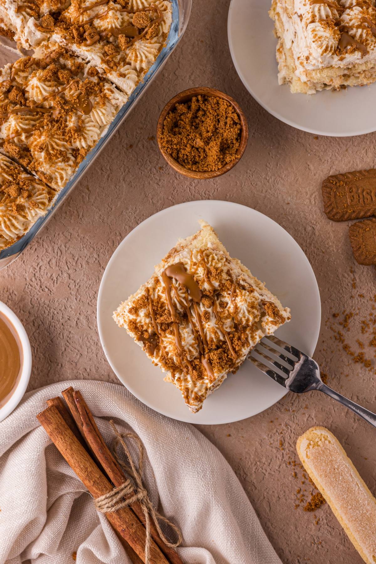 Overhead view of a plate with a slice of cake topped with cream and crumbs, surrounded by cinnamon sticks and brown sugar.