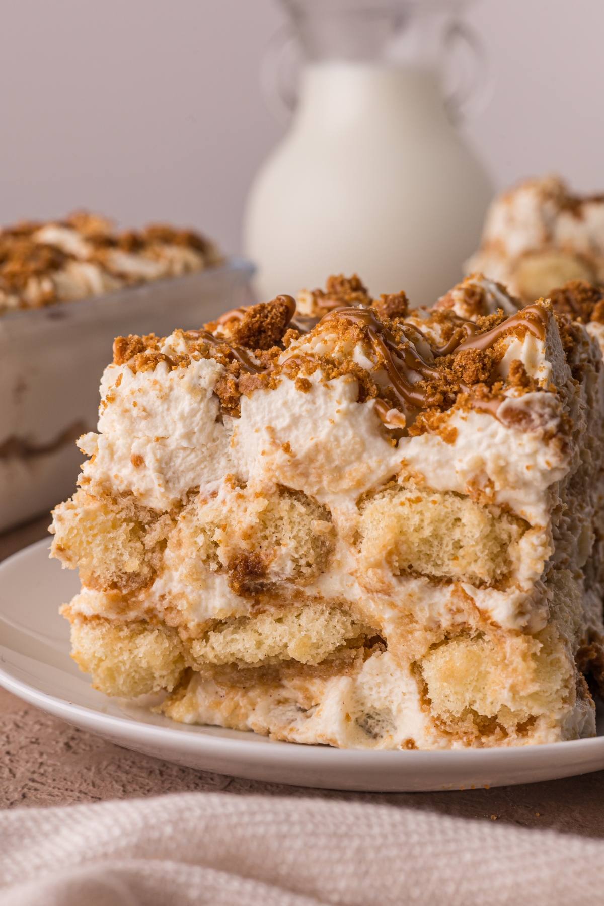 A close-up of a slice of creamy Biscoff tiramisu dessert on a white plate, topped with crushed cookies and caramel.