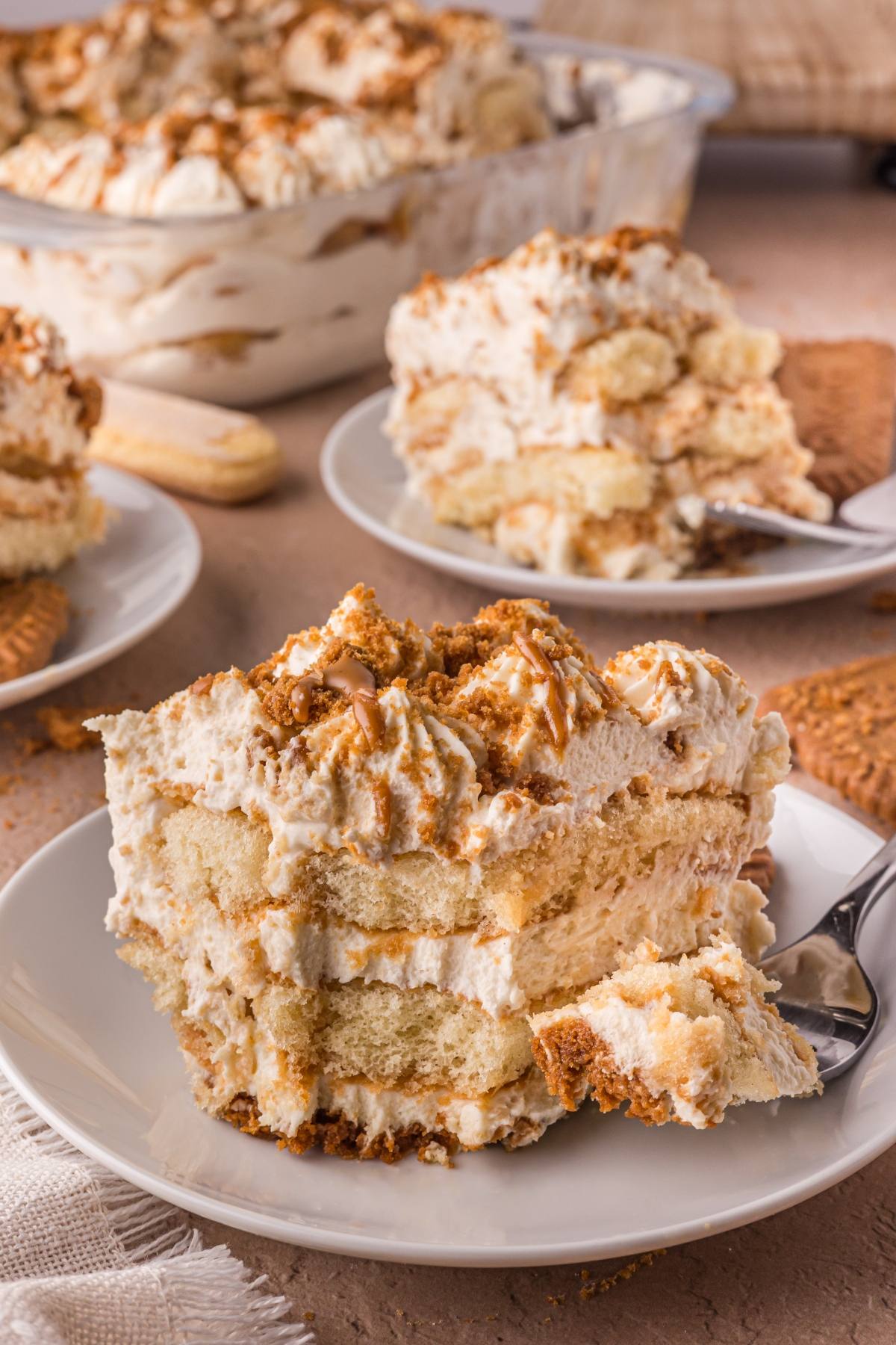A slice of creamy Biscoff tiramisu with layers of cake and cream on a white plate, topped with crumbs and caramel.