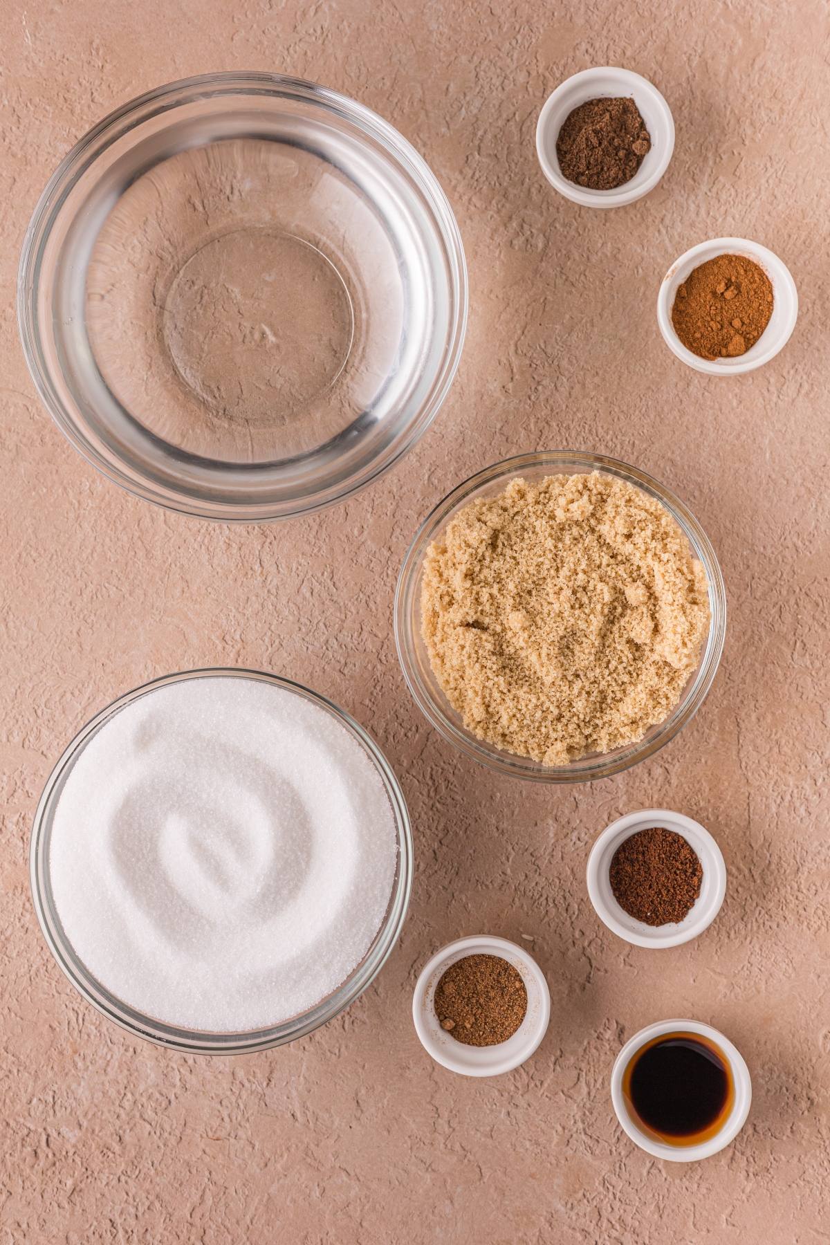 Overhead view of clear bowls with sugar, brown sugar, vanilla, and spices on a tan surface.
