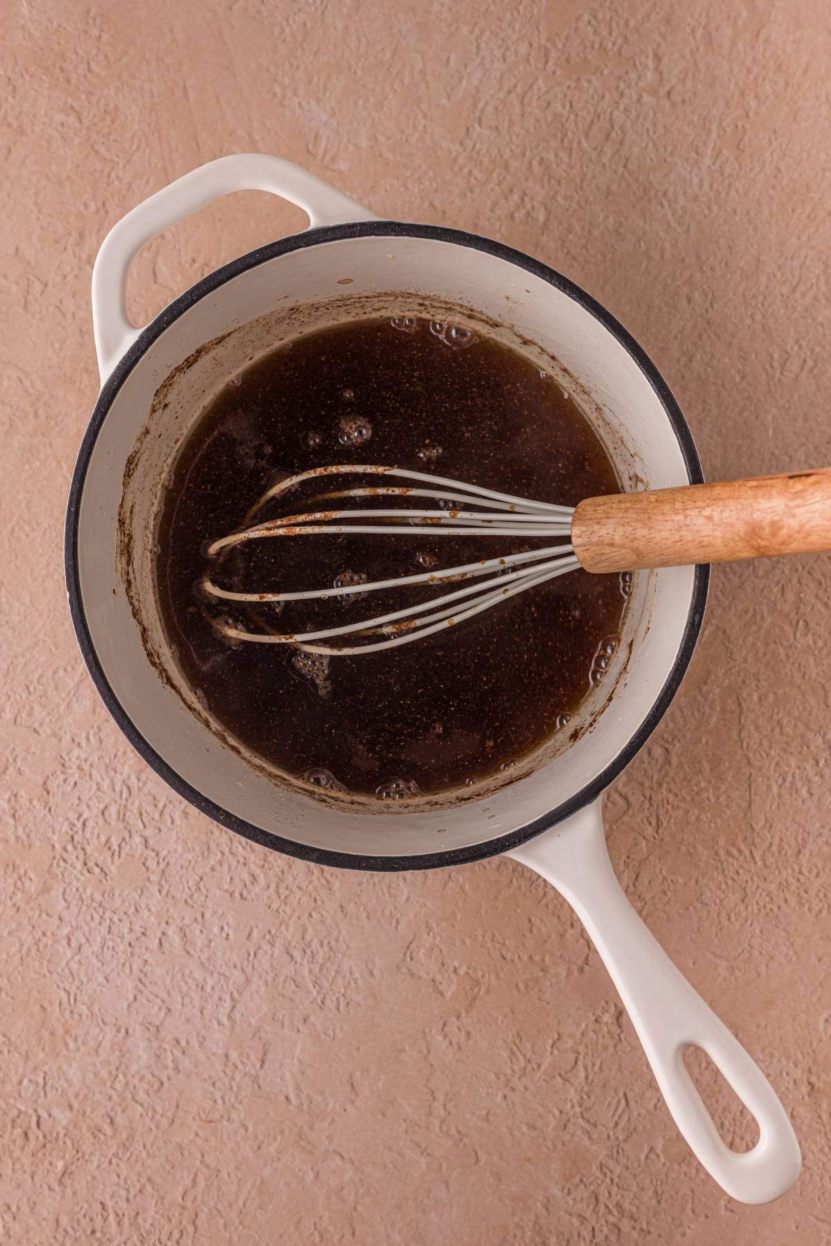 A saucepan with a whisk stirring a dark liquid on a light textured surface.