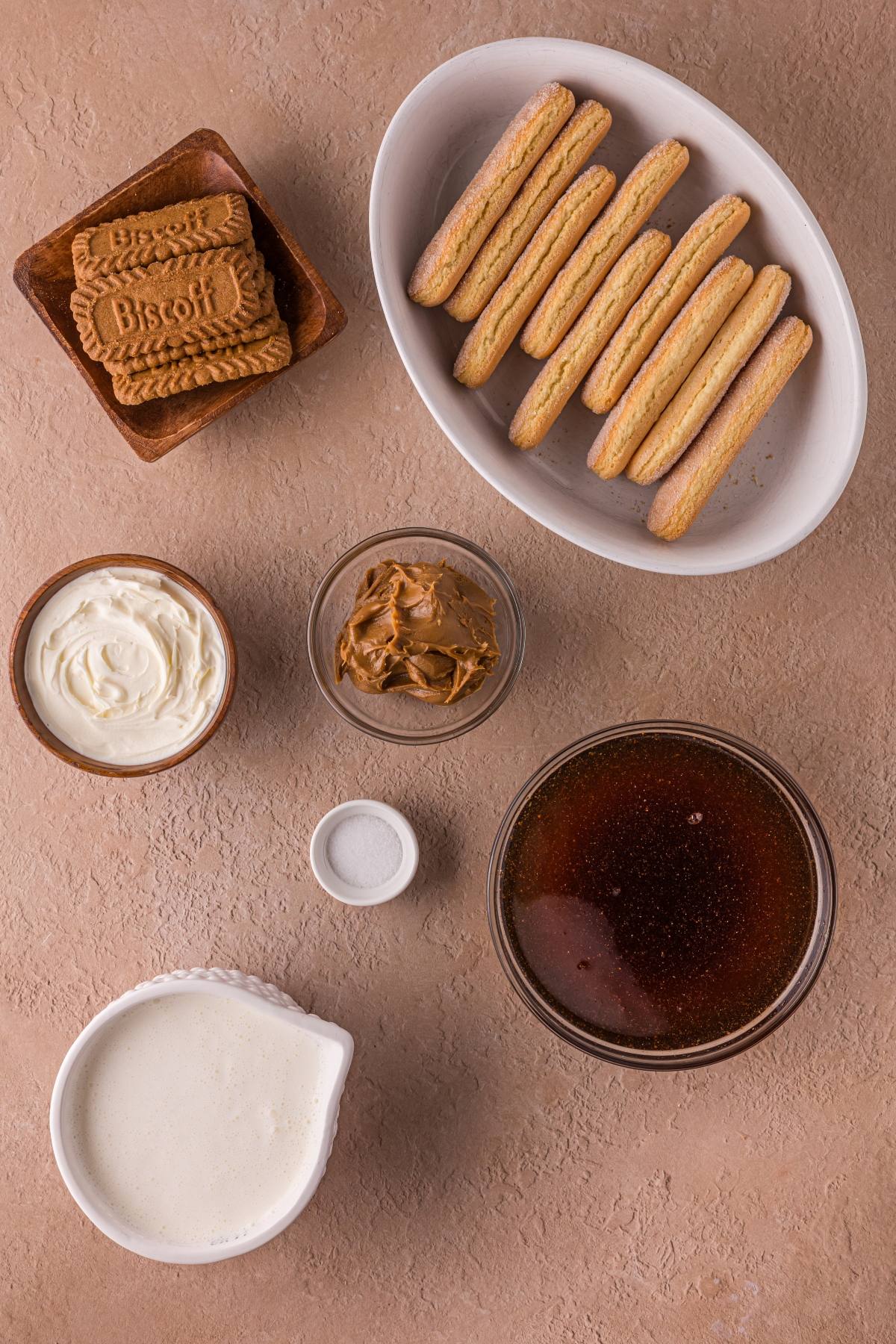 Top view of Biscoff cookies, ladyfingers, cream, peanut butter, coffee, salt, and whipped cream on a tan surface.