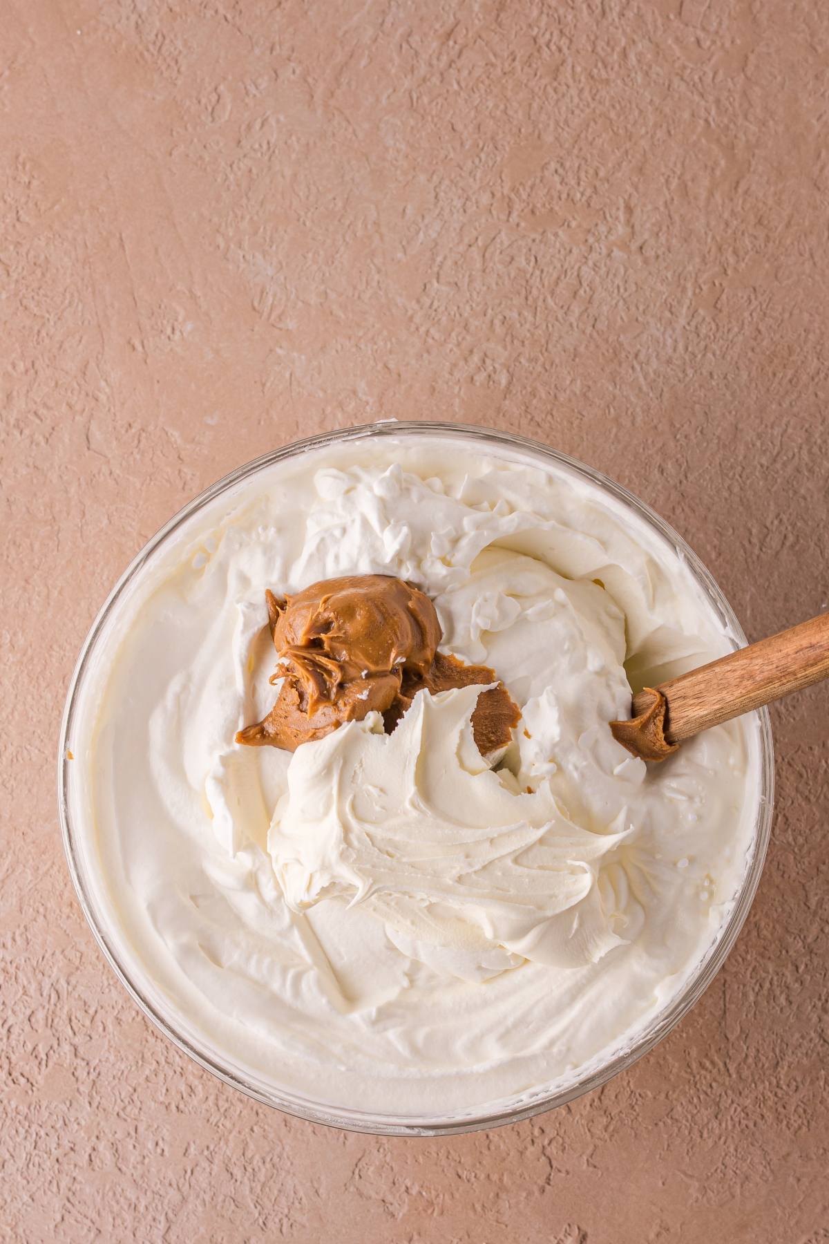 A glass bowl of whipped cream with a dollop of peanut butter and a wooden spoon on a textured surface.