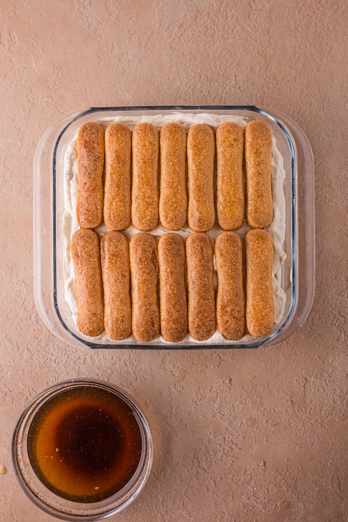 A glass dish with a layer of ladyfingers on cream, next to a bowl of coffee on a tan surface.